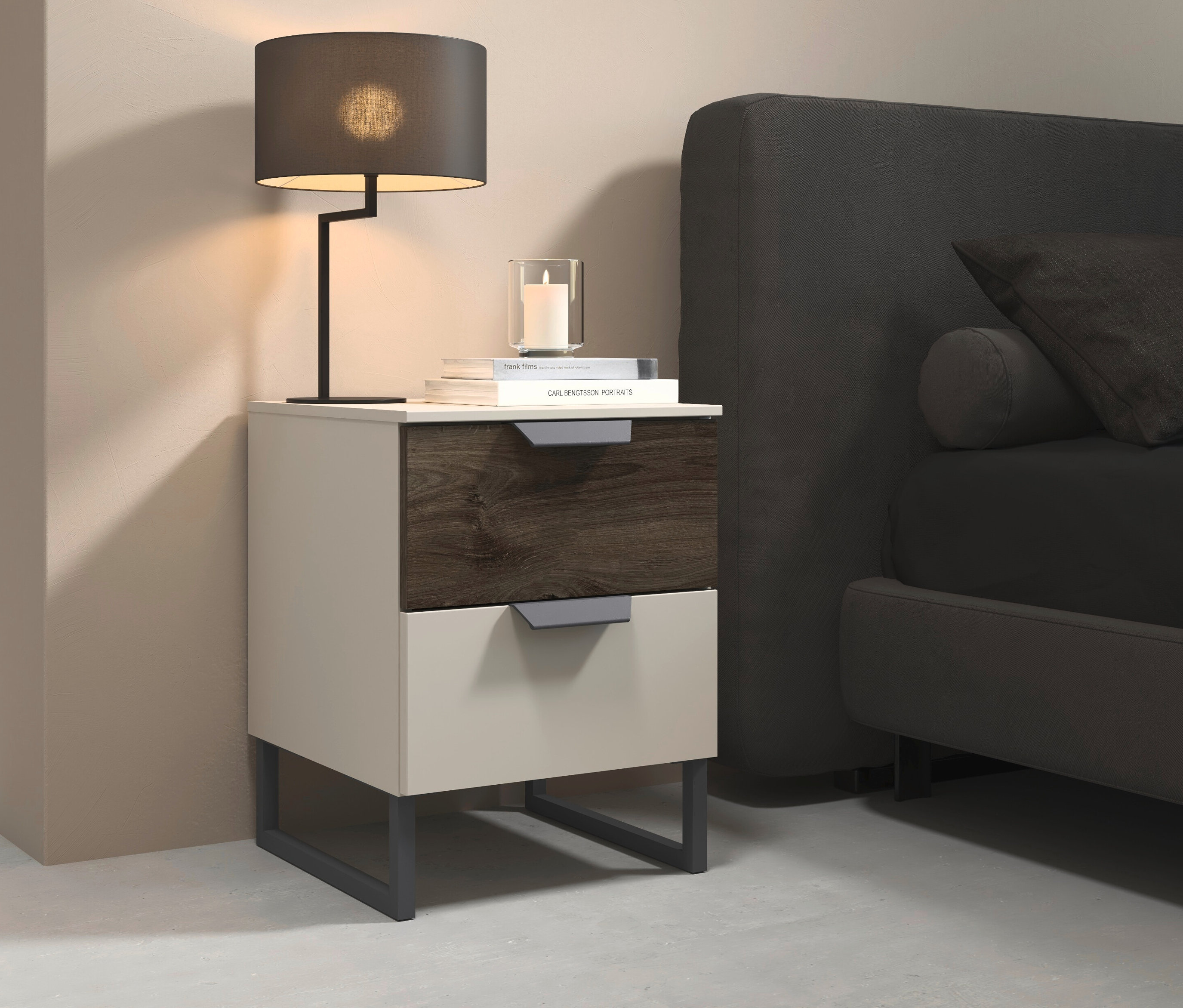 Auf einem Nachttisch mit zwei Schubladen stehen eine Lampe, ein Buch und eine Kerze. Daneben steht ein graues Bett.