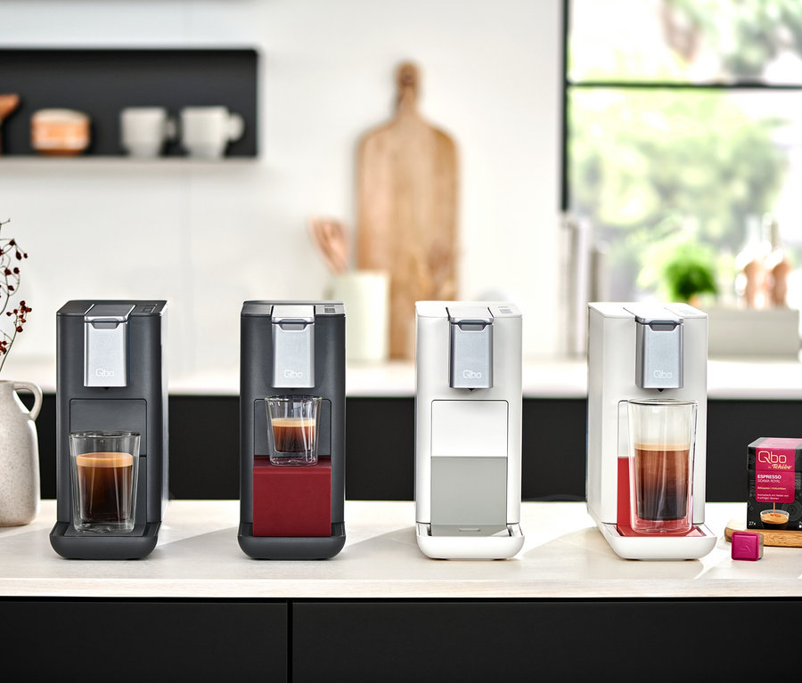 Vier Qbo ESSENTIAL Kaffeemaschinen in Dark Berry, Dark Stone, Bright Coral und Bright Sand stehen mit Espresso auf einer Küchentheke.