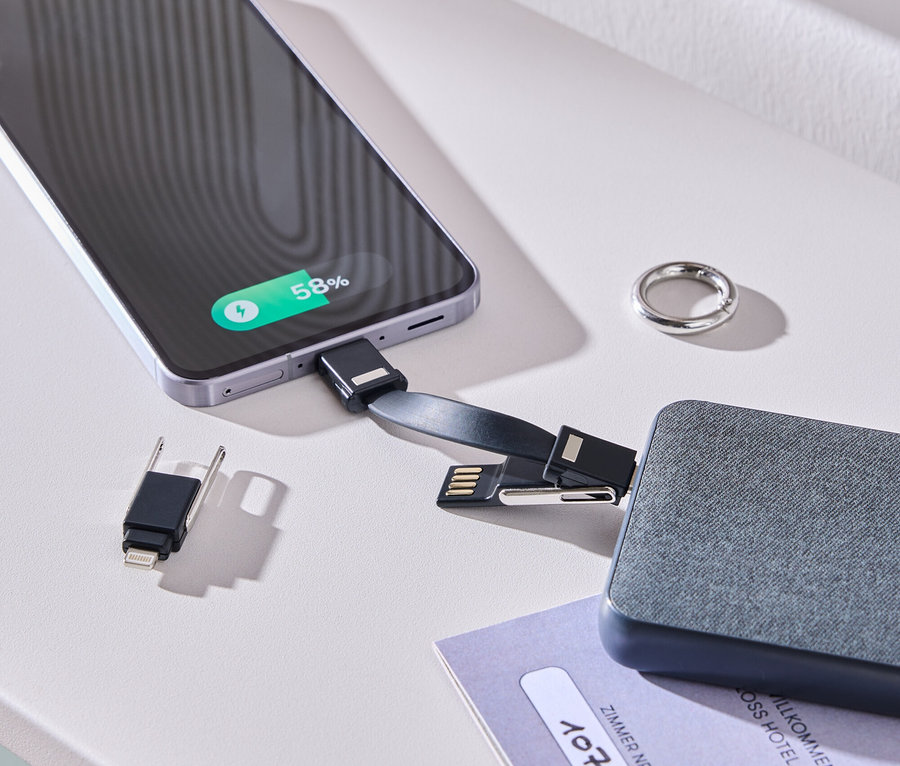 Ein Smartphone wird mit einem Ladekabel und einem grauen Akku auf einem weißen Tisch geladen. Ein Ring und eine Zimmerkarte liegen daneben.