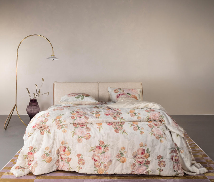 Essenza Satin-Bettwäsche »Orianna« in Normalgröße und Übergröße auf einem Bett mit beigem Kopfteil und einer goldenen Bogenlampe daneben.