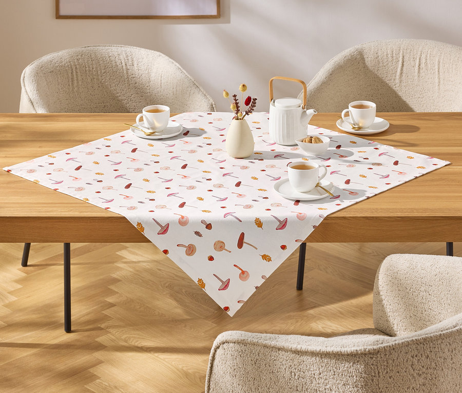 Auf einem Holztisch liegt eine Tischdecke mit Pilz- und Blättermuster, darauf Teetassen, eine Teekanne und eine Vase mit Blumen.
