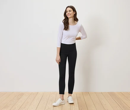 Eine Frau mit langen, braunen Haaren steht vor einer weißen Wand und trägt ein weißes Oberteil, schwarze Leggings und weiße Turnschuhe.