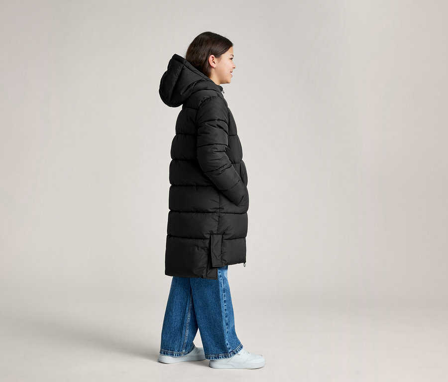 Eine Person trägt einen langen schwarzen Kapuzen-Steppmantel, blaue Jeans und weiße Turnschuhe und steht im Profil.