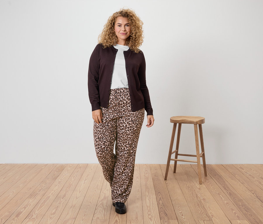 Frau mit lockigem Haar trägt eine braune Strickjacke, ein weißes T-Shirt und eine Hose mit Leopardenmuster.