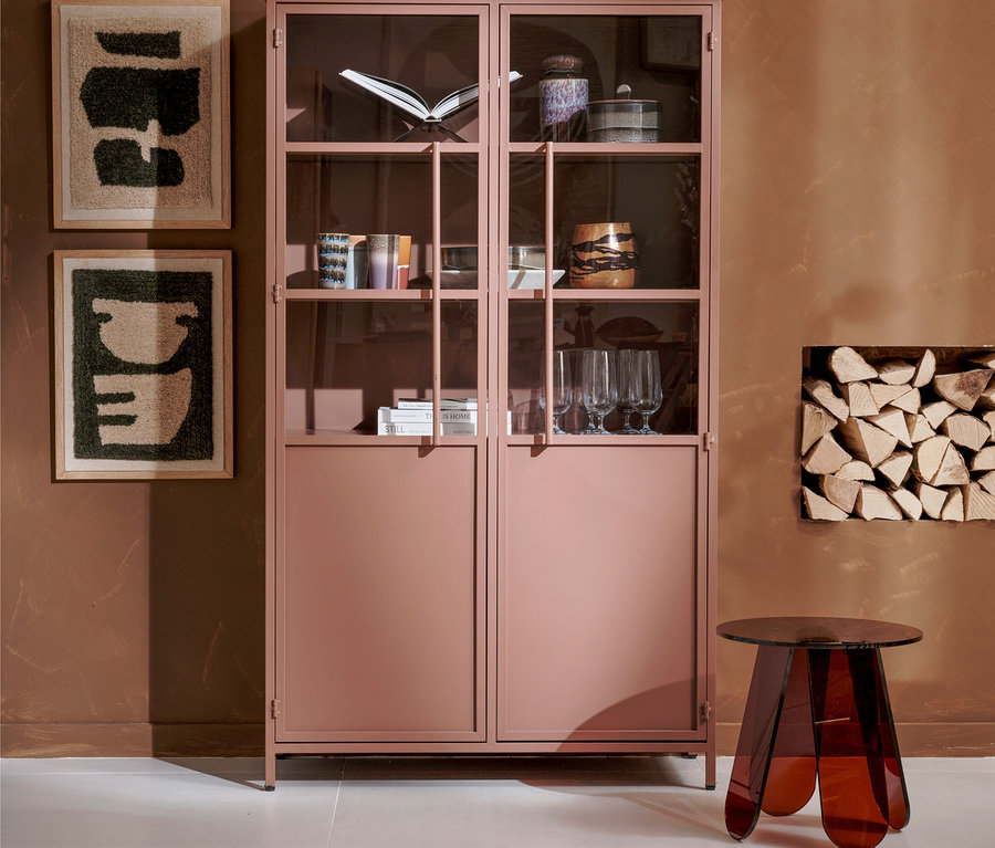 Ein rosa Metallschrank mit Glasvitrine, gefüllt mit Büchern und Gläsern, steht vor einer braunen Wand. Links hängen zwei abstrakte Bilder, rechts ein roter Beistelltisch und ein Fach mit Brennholz.