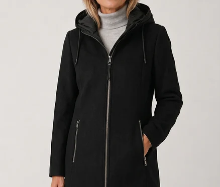 Frau in schwarzem Mantel mit Kapuze und grauem Rollkragenpullover, lächelt in die Kamera.