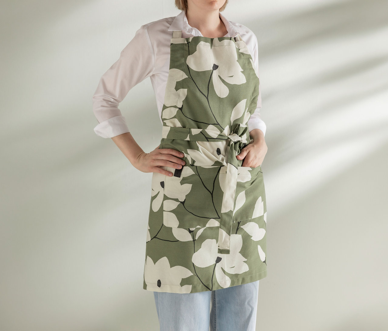 Eine Person trägt ein weißes Hemd, hellblaue Jeans und eine grüne Schürze mit weißem Blumenmuster. Eine Hand steckt in einer Schürzentasche, die andere ist in die Hüfte gestemmt, vor einer hellen Wand mit Schatten.