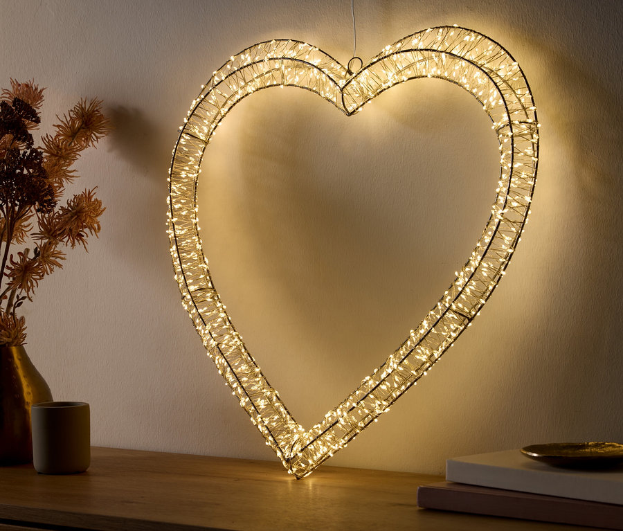 Ein großes, herzförmiges Lichtobjekt aus Draht, umwickelt mit warmweißen LED-Lichtern, leuchtet hell. Es steht auf einer Holzoberfläche und lehnt an einer hellen Wand. Links davon steht eine goldene Vase mit Trockenblumen und ein kleiner grauer Topf. Rechts davon liegen zwei gestapelte Bücher mit einer kleinen goldenen Schale darauf.