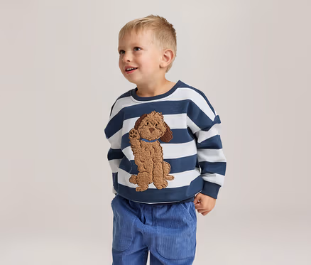 Ein kleiner Junge steht lächelnd und blickt nach oben. Er trägt ein weiß-blau gestreiftes Sweatshirt mit einer braunen Hundeapplikation und eine blaue Cordhose.