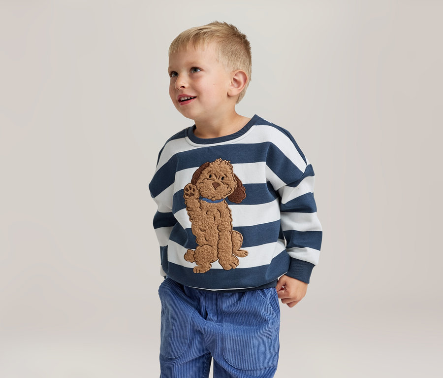 Ein kleiner Junge steht lächelnd und blickt nach oben. Er trägt ein weiß-blau gestreiftes Sweatshirt mit einer braunen Hundeapplikation und eine blaue Cordhose.
