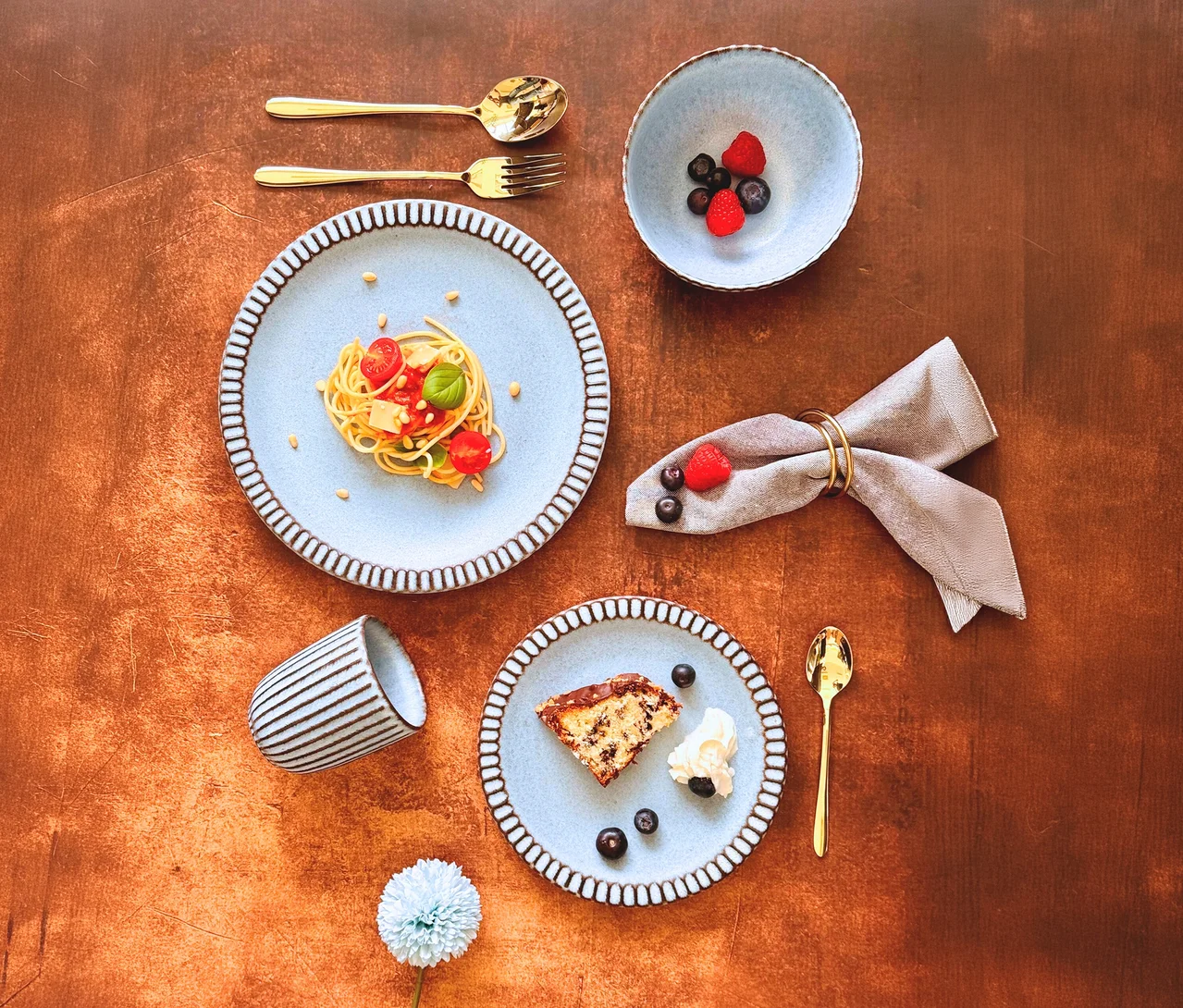 Draufsicht auf einen rustikalen Tisch mit hellblauen Keramiktellern mit gewelltem Rand, die Pasta und Kuchen mit Beeren servieren. Dazu goldfarbenes Besteck vom Mäser Besteck-Set »Ebina«, eine gestreifte Tasse und eine Serviette.
