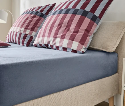 Ein Bett mit beigem, gepolstertem Kopfteil und Rahmen, bezogen mit einem blaugrauen Spannbetttuch. Zwei große Kissen und ein kleines Kissen mit rot-weiß-blauem Karomuster liegen auf dem Bett. Eine weiße Lampe und ein Nachttisch sind im Hintergrund sichtbar.