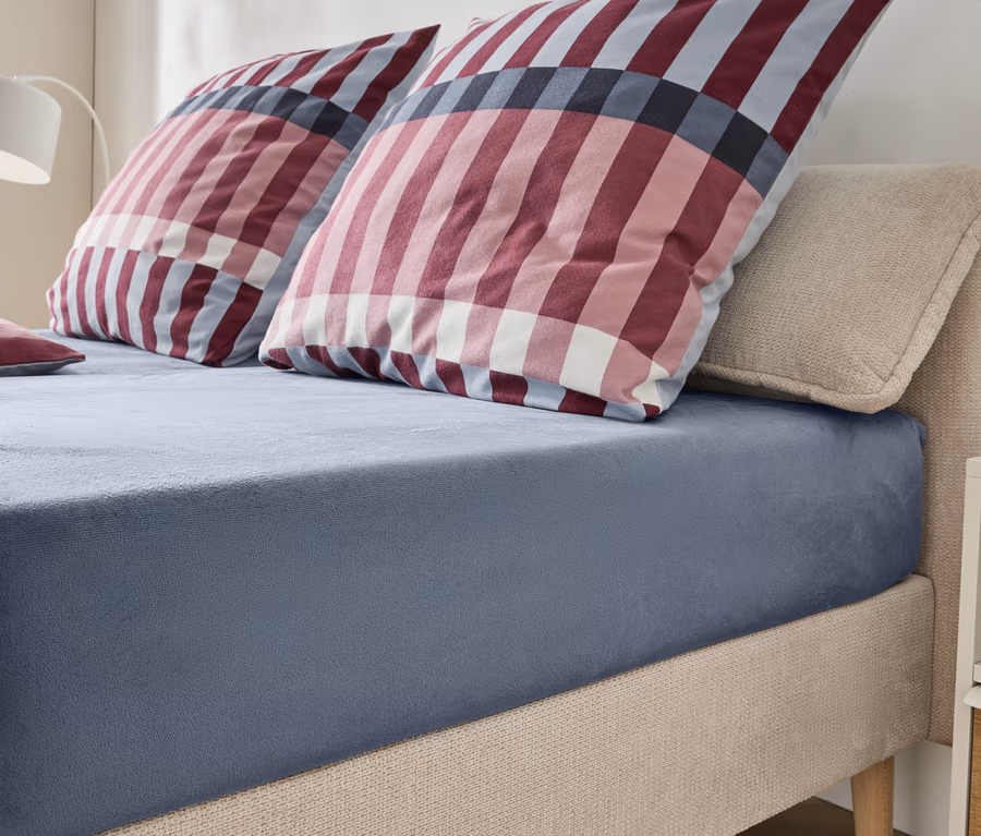 Ein Bett mit beigem, gepolstertem Kopfteil und Rahmen, bezogen mit einem blaugrauen Spannbetttuch. Zwei große Kissen und ein kleines Kissen mit rot-weiß-blauem Karomuster liegen auf dem Bett. Eine weiße Lampe und ein Nachttisch sind im Hintergrund sichtbar.
