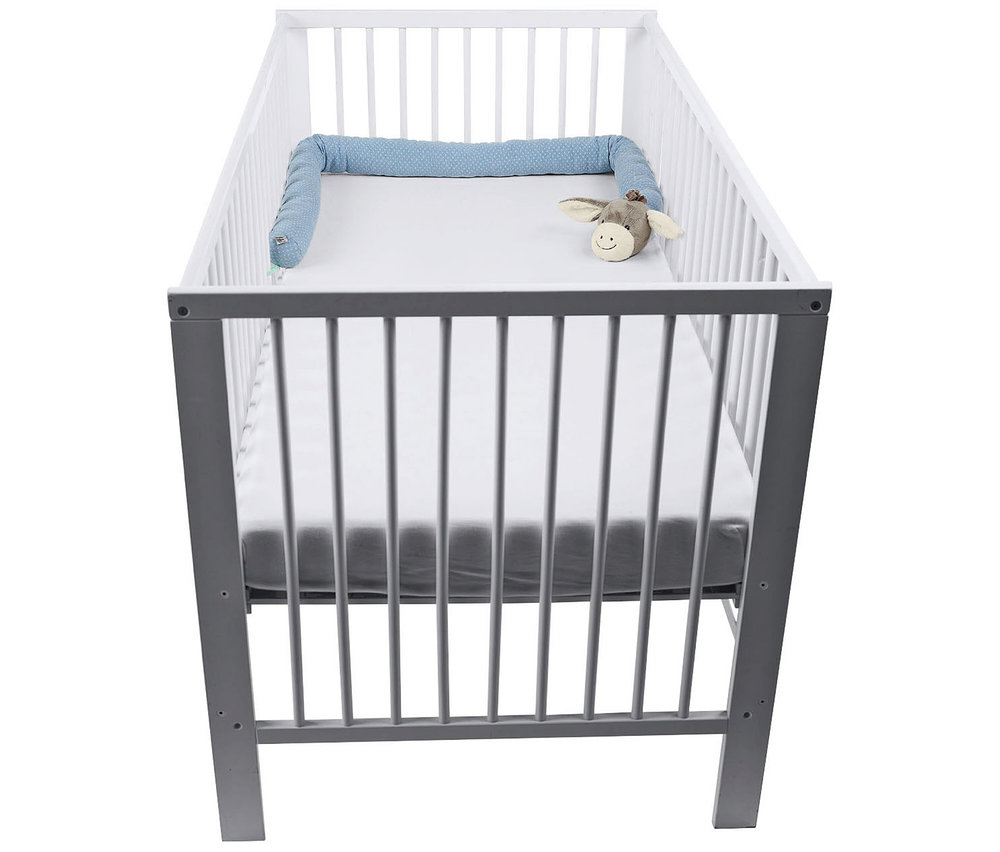 Weißes und graues Babybett mit weißer Matratze, einer blau gemusterten Bettschlange an einer Seite und einem kleinen Plüsch-Esel am Kopfende.