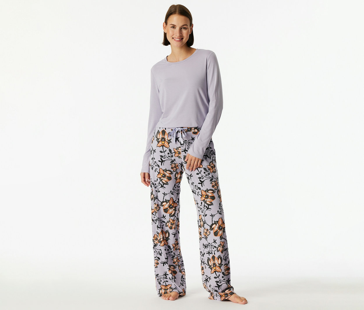 Lächelnde Frau steht in einem hellvioletten Langarmshirt und einer hellvioletten Pyjamahose mit orange-schwarzem Blumenmuster.