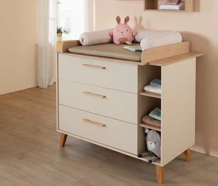 Eine hellbeige Wickelkommode mit drei Schubladen und zwei offenen Regalfächern. Oben liegen eine Wickelauflage, ein rosafarbenes Hasen-Stofftier und ein Kissen. Gefaltete Babykleidung liegt in den Fächern, ein graues Bären-Stofftier lugt aus dem untersten Fach. Im Hintergrund ist ein Fenster mit einem transparenten Vorhang zu sehen.