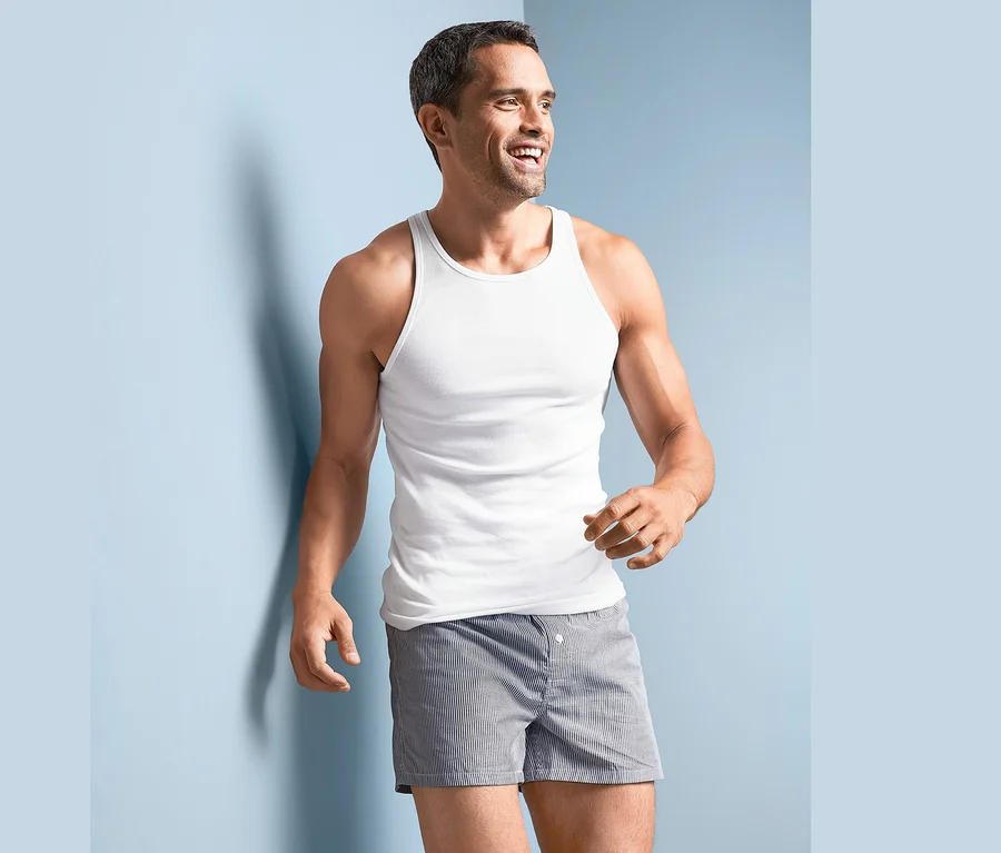 Ein lächelnder Mann in einem weißen 2 Feinripp-Unterhemd und gestreiften Shorts steht an einer blauen Wand.