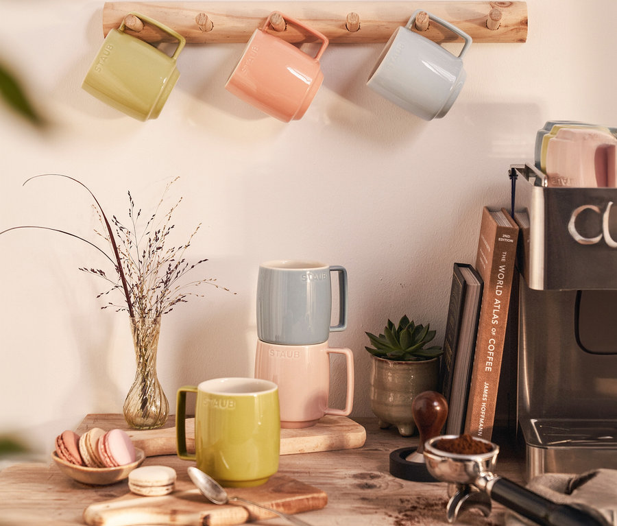 Mehrere STAUB Kaffeebecher »Macaron« in verschiedenen Farben hängen an einer Holzleiste und stehen auf einem Holztisch.