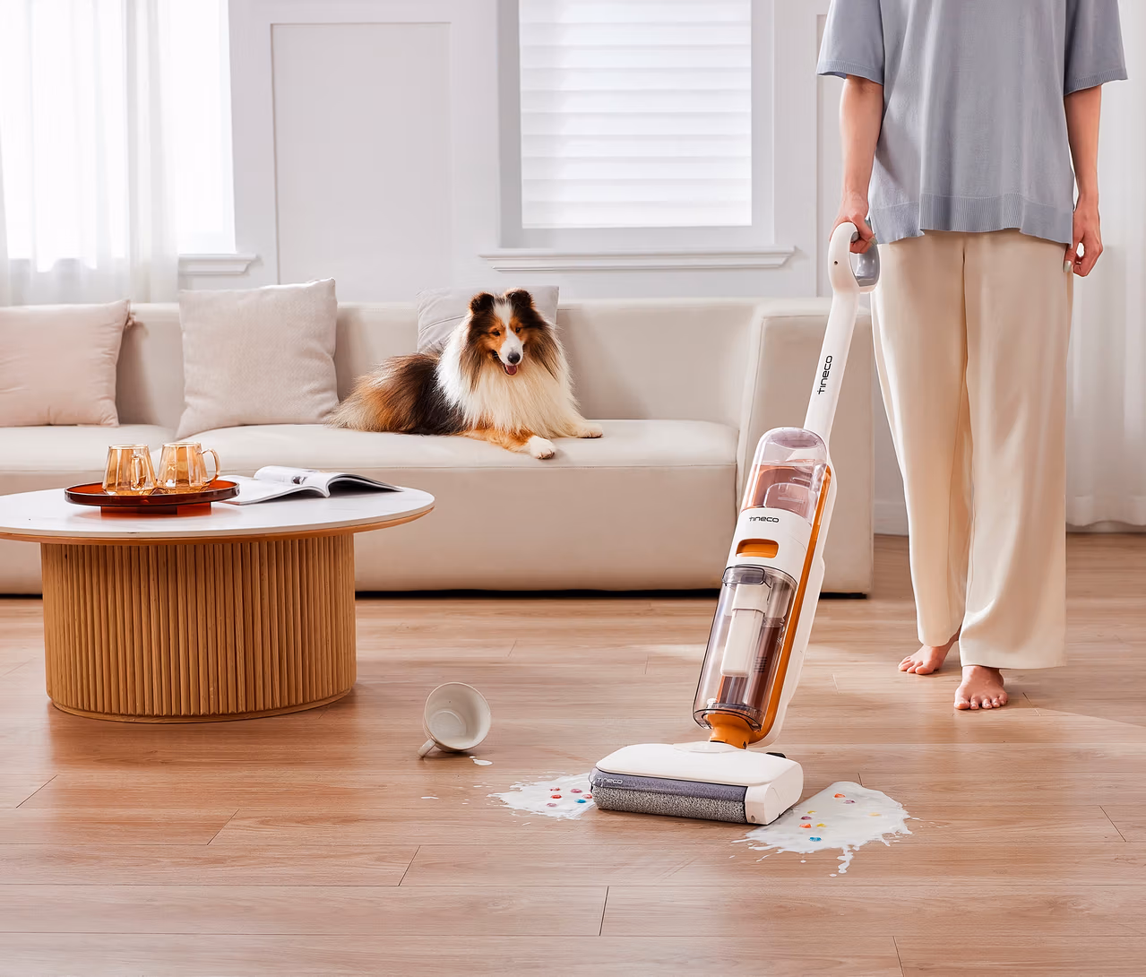 Eine Person reinigt mit einem weiß-orangen Tineco 2-in-1-Nass-Trockensauger »Y2 Plus« eine verschüttete Flüssigkeit mit bunten Streuseln auf einem Holzfußboden. Eine umgekippte weiße Tasse liegt daneben. Im Hintergrund liegt ein flauschiger braun-weißer Hund auf einem hellen Sofa neben einem Couchtisch mit Gläsern und einem Buch.