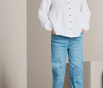 Zwei Kinder posieren auf grauen Blöcken. Das Mädchen steht und trägt eine weiße Bluse und Schlupf-Denim. Der Junge sitzt und trägt einen gestreiften Pullover und Schlupf-Denim.