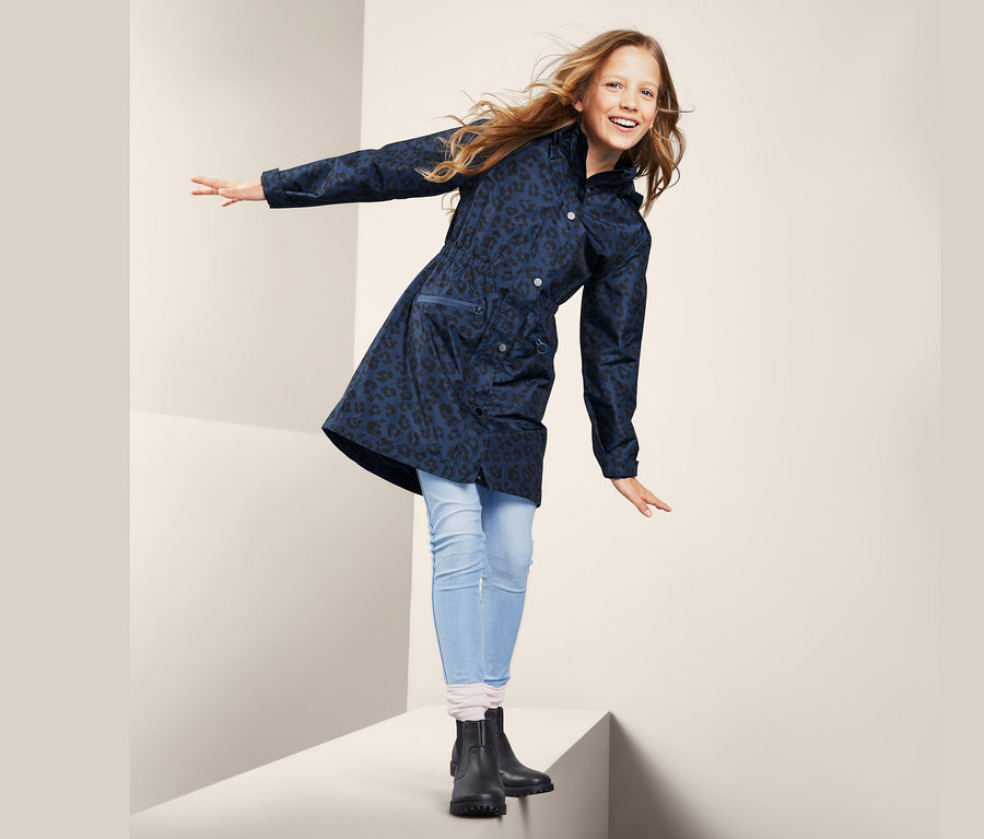 Mädchen steht auf einem weißen Würfel und winkt mit den Händen. Sie trägt einen Allwetterparka mit Alloverprint, Jeans und schwarze Stiefel.