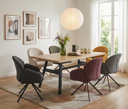 Ein modernes Esszimmer mit einem Holztisch und sechs Stühlen in verschiedenen Farben auf einem Teppich. Über dem Tisch hängt eine große, runde Papierlampe, und an der Wand sind vier gerahmte abstrakte Bilder zu sehen.