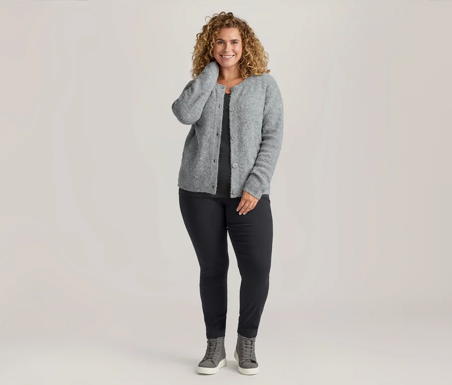 Lächelnde Frau in grauem Bouclé-Strickcardigan, dunklem Langarm-Shirt und schwarzer Stretchhose, dazu graue Sneaker.