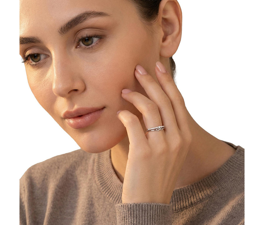 Frau mit hellem Teint und natürlichem Make-up, die einen hellbraunen Pullover trägt. Am Ringfinger ihrer rechten Hand, die ihre Wange berührt, trägt sie einen silberfarbenen Amor Ring mit Infinity-Symbol und kleinen Steinen.
