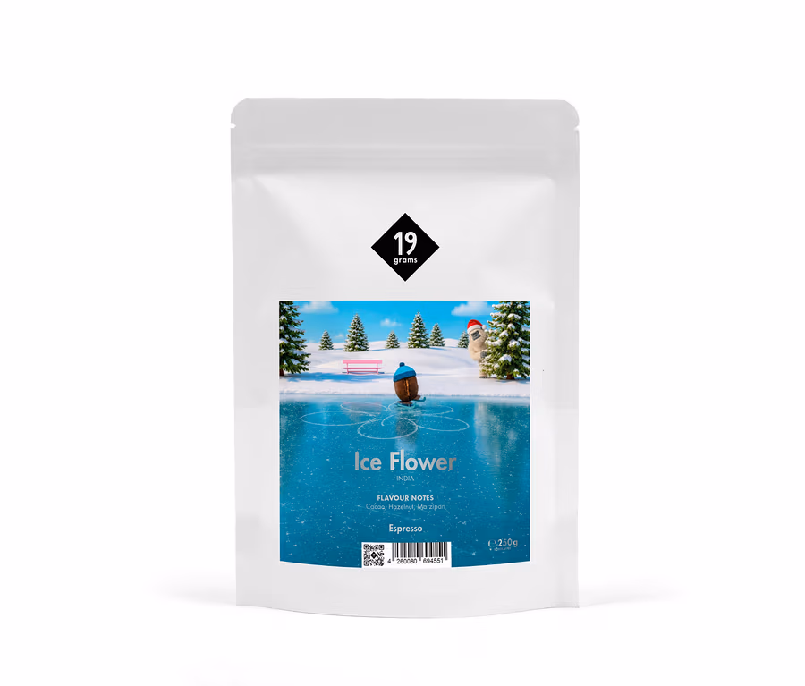 Weiße Kaffeeverpackung mit dem Logo „19 grams“ und einer Illustration einer Winterlandschaft mit einer Figur, die auf einer Eisfläche Schlittschuh läuft, umgeben von verschneiten Bäumen und einer Bank. Darunter steht „Ice Flower“, „INDIA“, „FLAVOUR NOTES Cacao, Hazelnut, Marzipan“, „Espresso“ und „250g“.