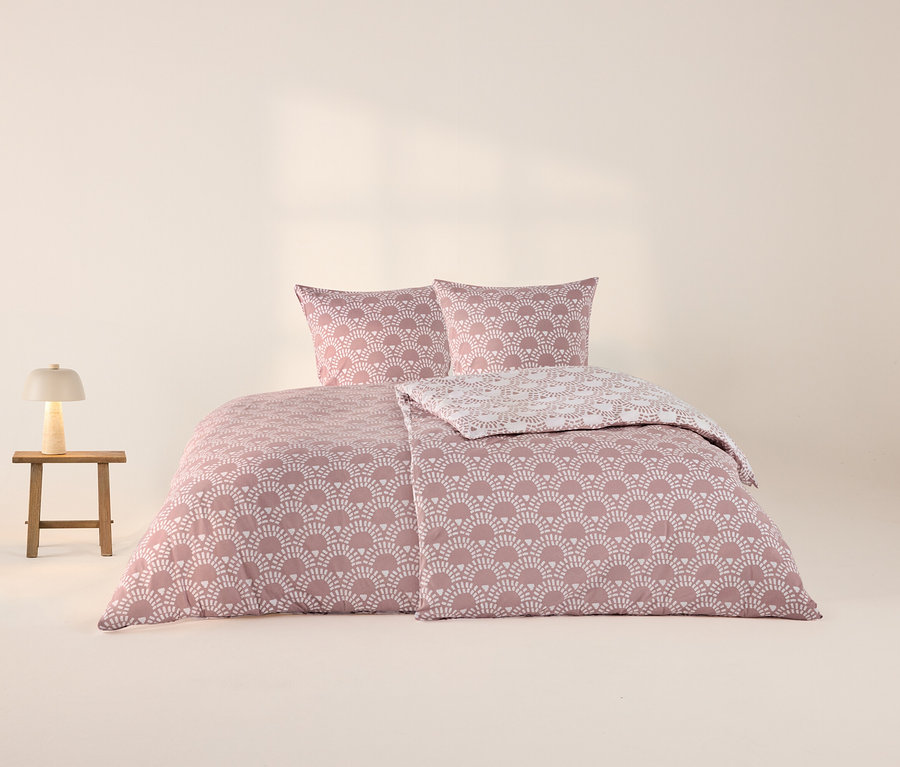 Rosa Jersey-Bettwäsche mit weißem Fächermuster auf einem Bett, mit zwei Kissen und einer zurückgeschlagenen Decke. Daneben steht ein kleiner Holztisch mit einer leuchtenden weißen Pilzlampe.
