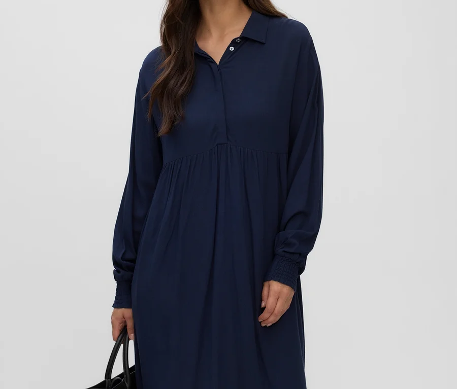 Frau mit langen dunklen Haaren, die ein dunkelblaues MAMALICIOUS Kleid »ESRA LIA« mit Kragen, Knöpfen und geraffter Taille trägt und eine schwarze Tasche hält.