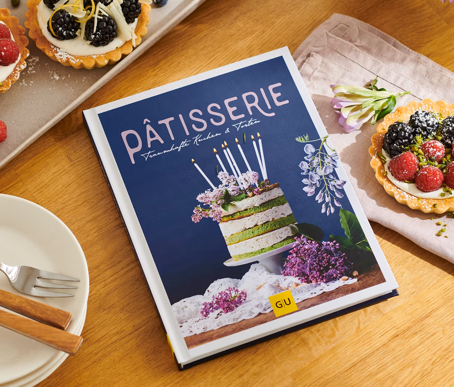 Auf einem Holztisch liegen ein Teller mit kleinen Törtchen, das Buch »Pâtisserie«, Teller und eine Serviette.