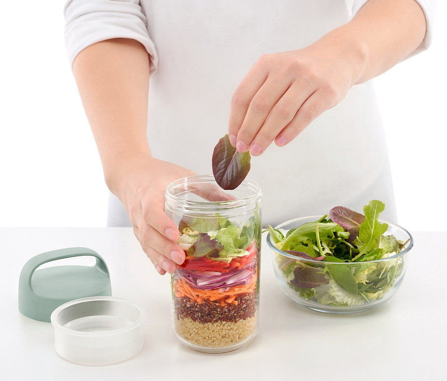 Person fügt Salatblatt in To-go-Lunchbox, daneben Schüssel mit Salat.