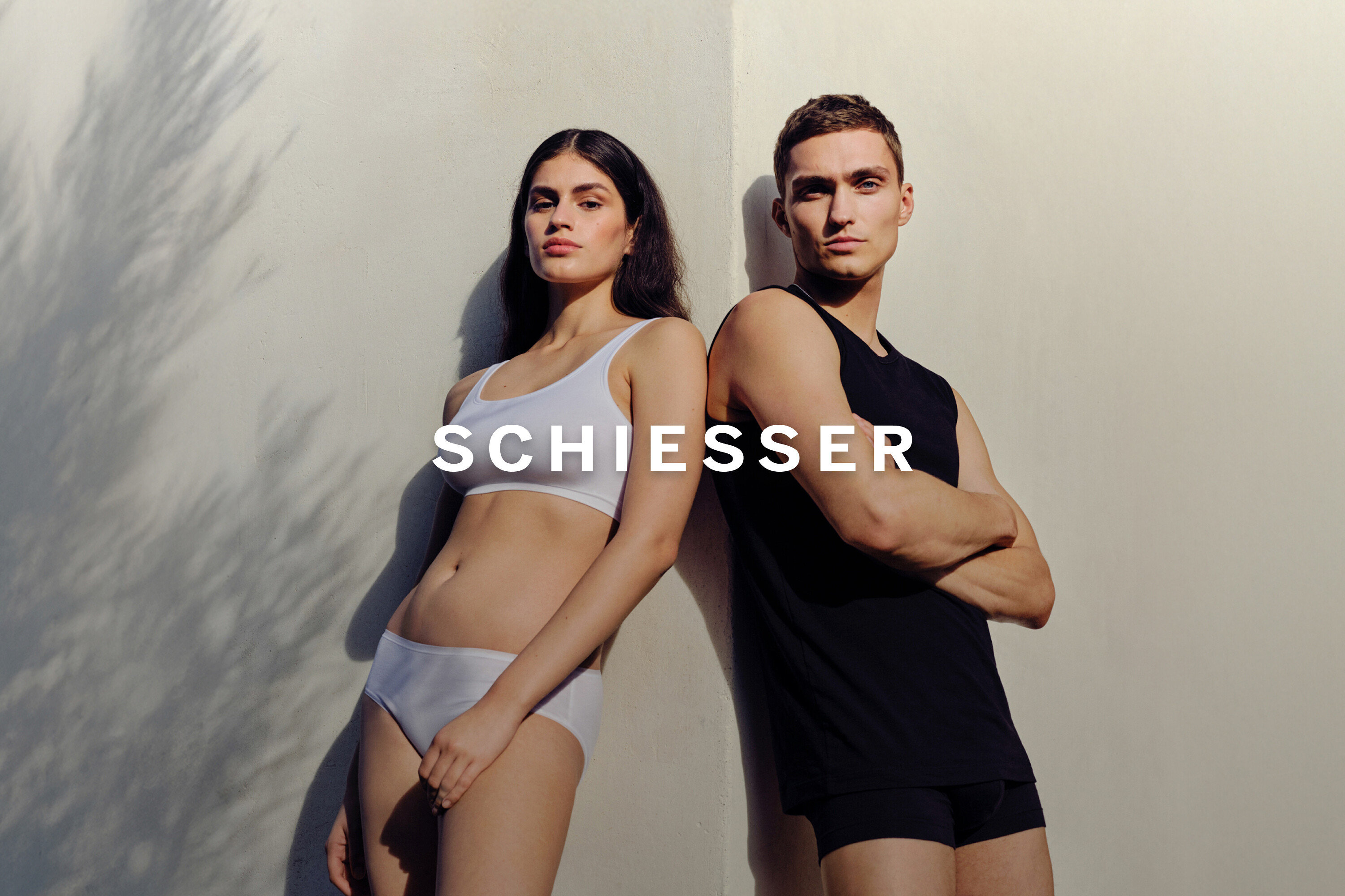 Frau in weißem Sport-BH und Unterhose lehnt an der Wand, neben einem Mann in schwarzem Tanktop und Unterhose.