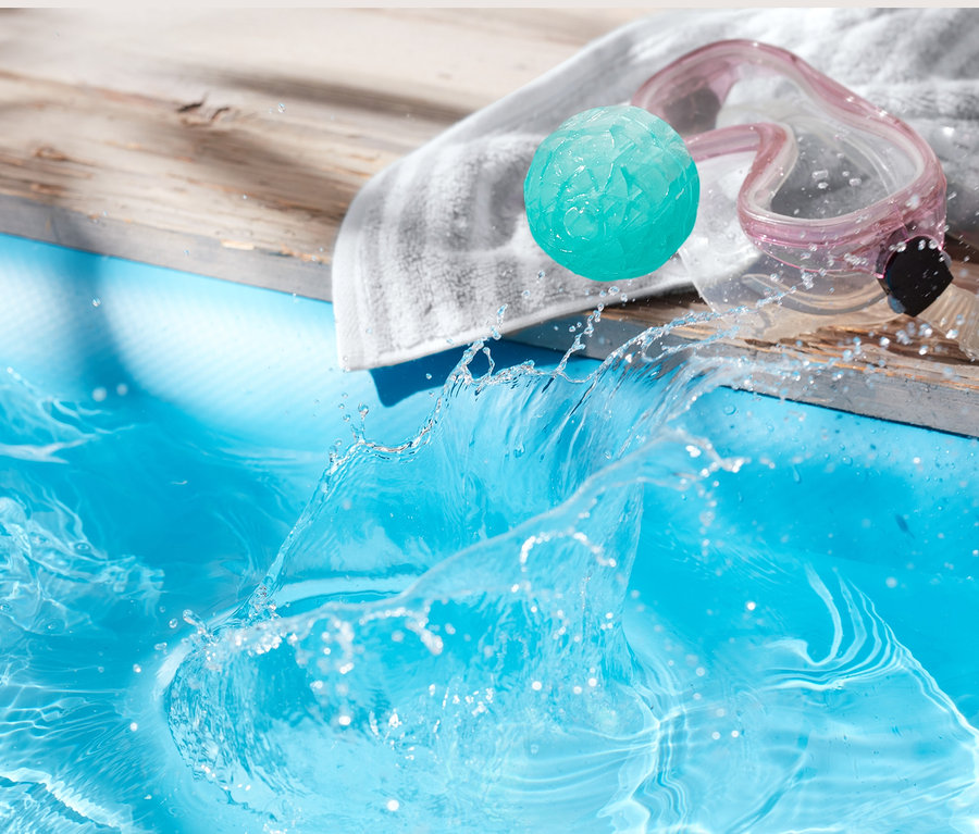 Ein hüpfender Wasserball liegt neben einer Brille auf einem Handtuch am Pool.