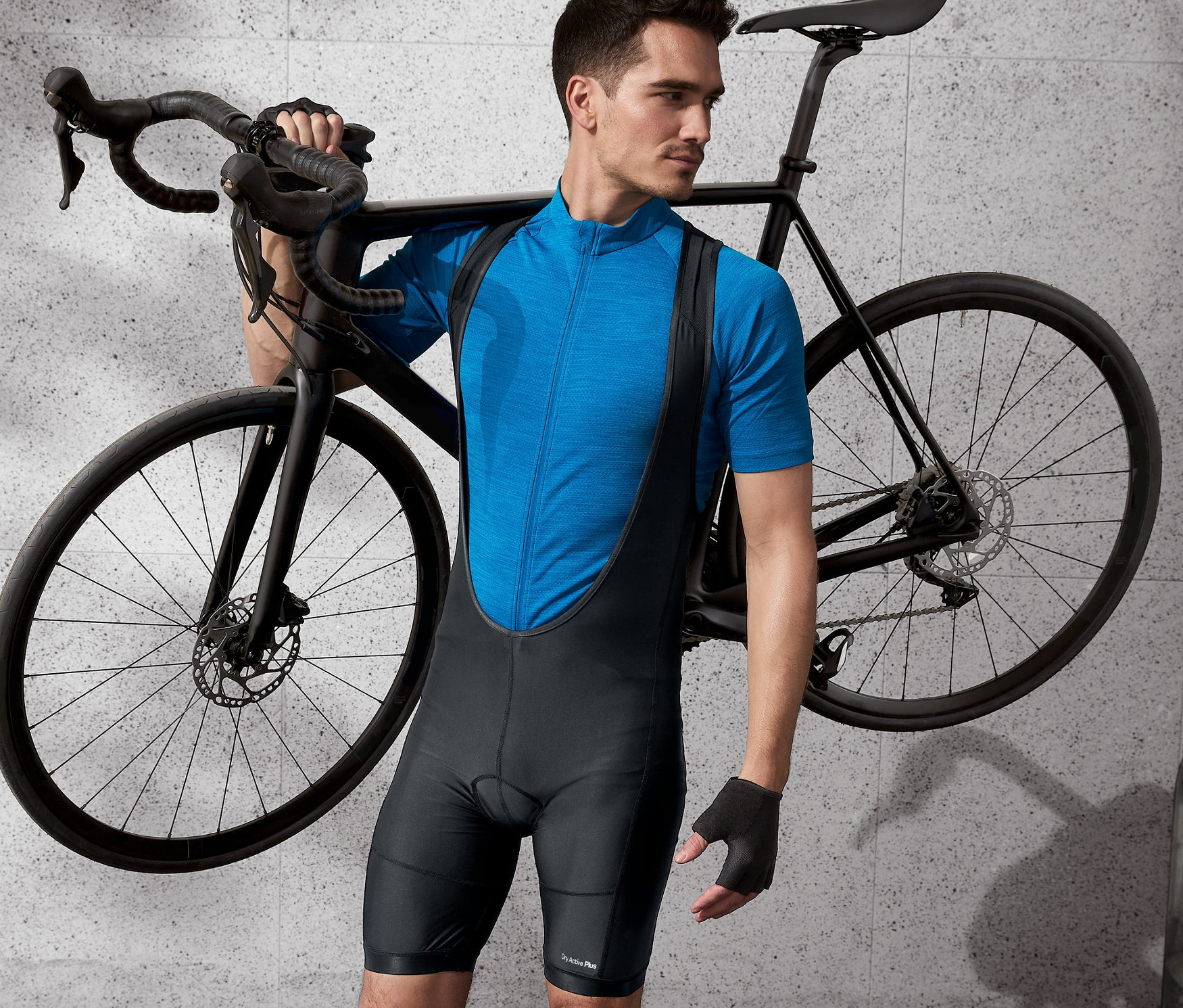 Mann in Fahrrad-Trägerhose und blauem Trikot trägt ein schwarzes Fahrrad über der Schulter.