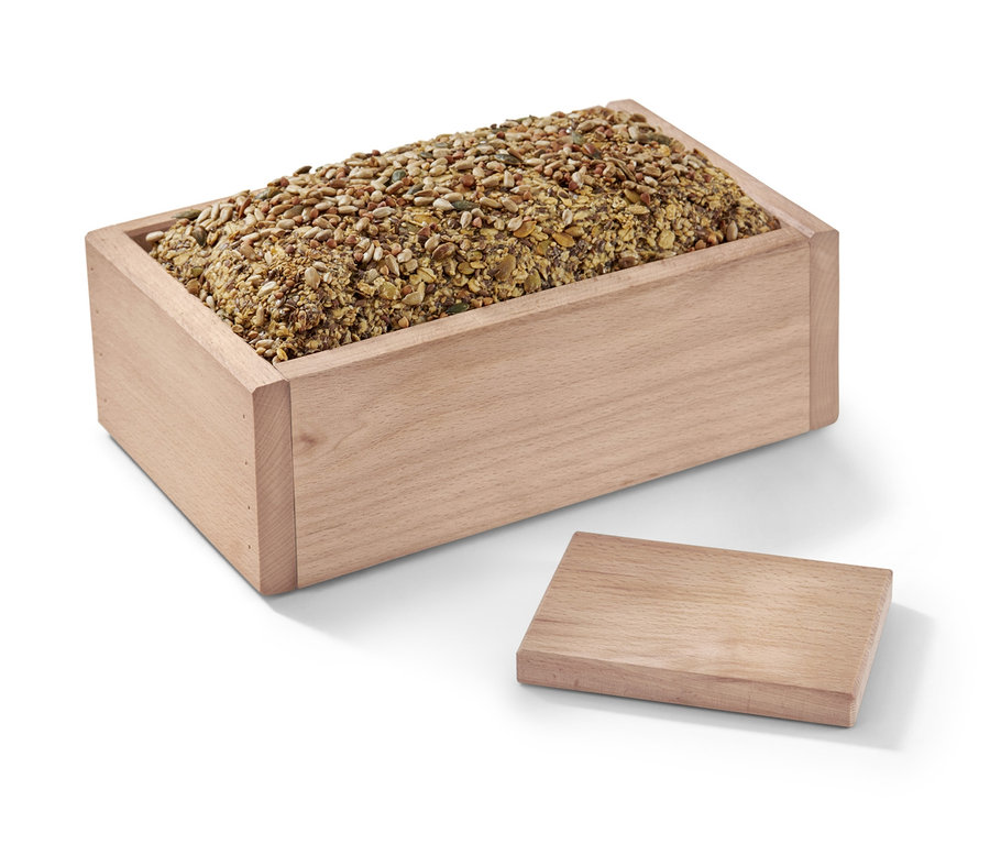Ein Holz-Brotbackrahmen gefüllt mit Körnern und Samen, daneben liegt ein Deckel.