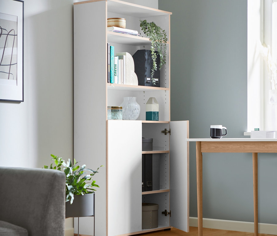 Ein hohes, halboffenes Regal mit Büchern und Dekorationen steht in einem Wohnzimmer.