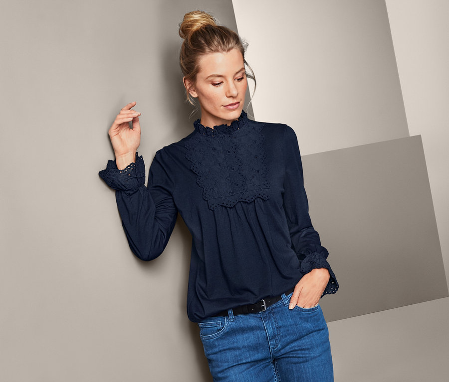 Frau, die sich an eine Wand lehnt, trägt ein Blusenshirt mit Stehkragen und Jeans.