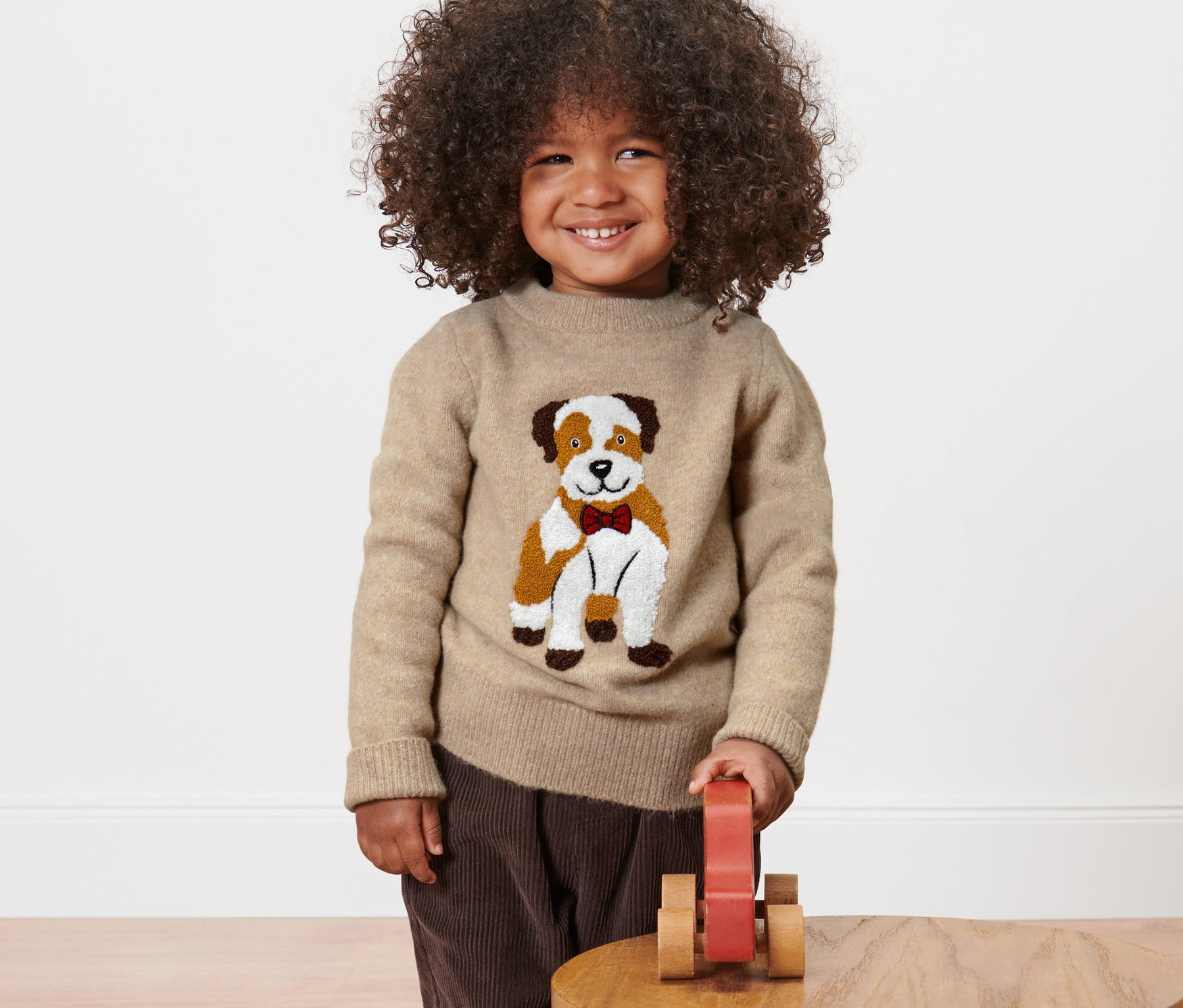 Ein lächelndes Kind mit lockigem Haar trägt einen Kleinkind-Strickpullover mit Applikation und spielt mit einem Holzspielzeug.