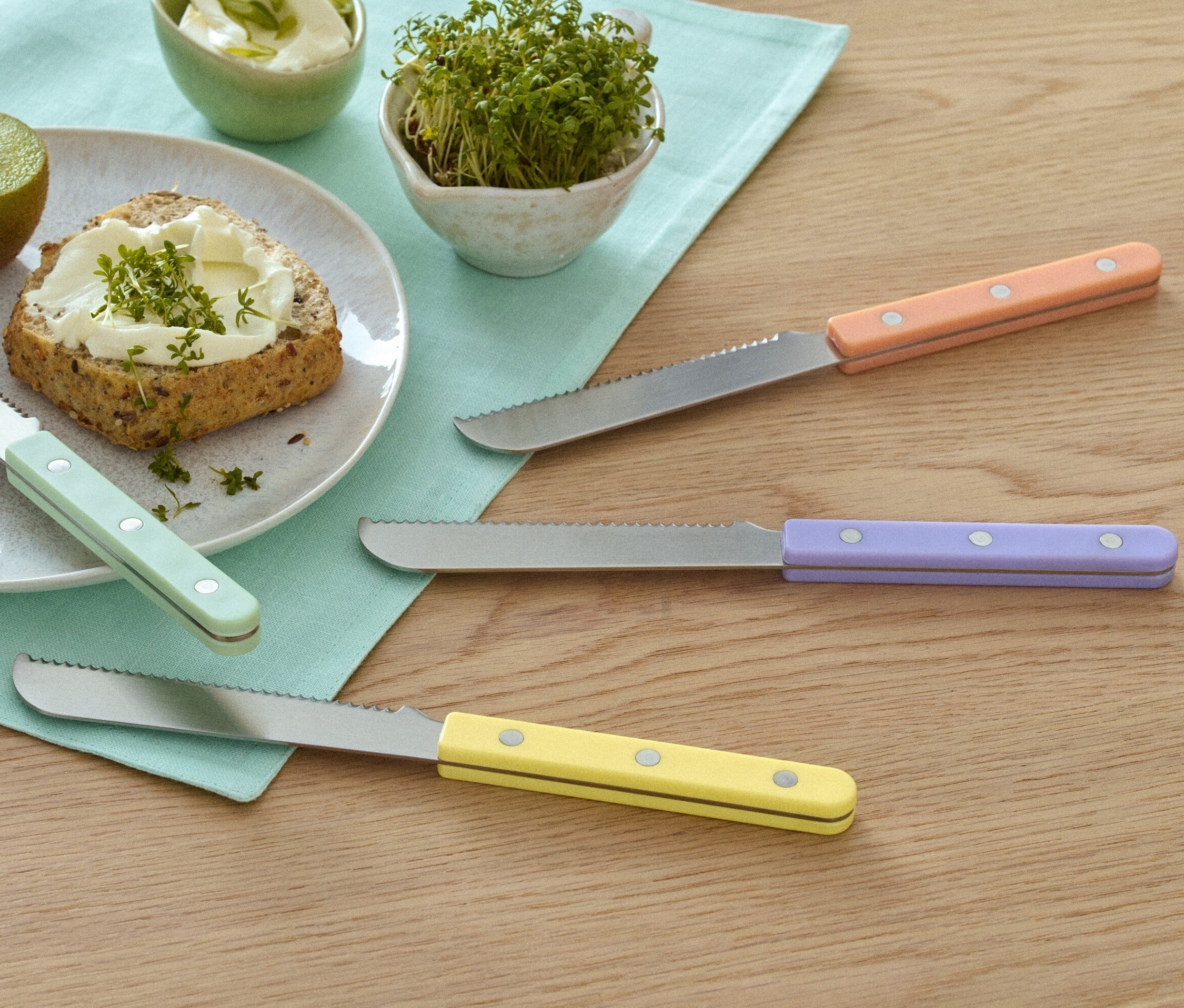 Teller mit Kiwi, Brot mit Aufstrich und Kresse neben 4 Frühstücksmessern.