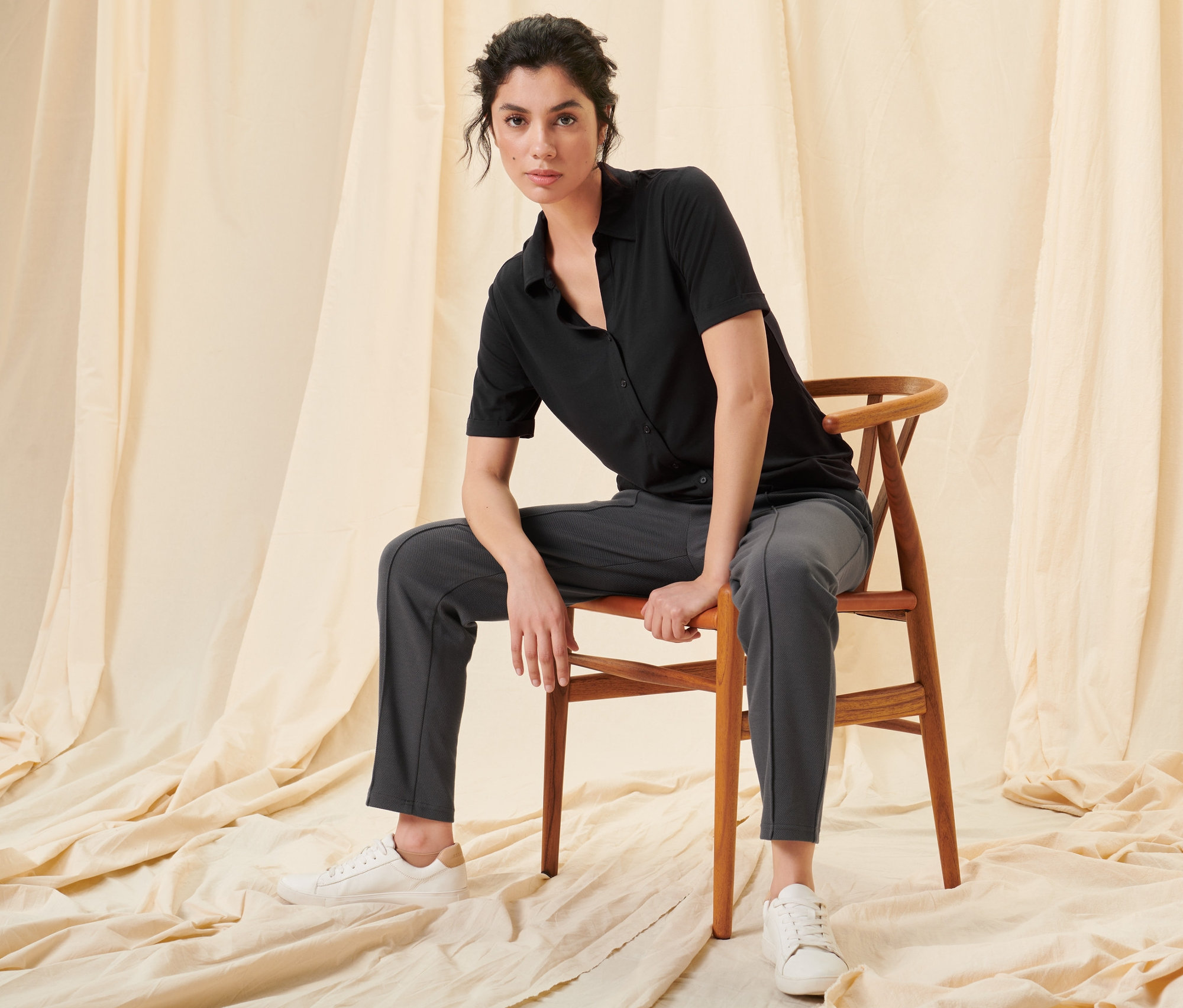 Frau sitzt auf einem Stuhl und trägt ein schwarzes Blusenshirt mit Knopfleiste und eine Joggpants mit Biese.