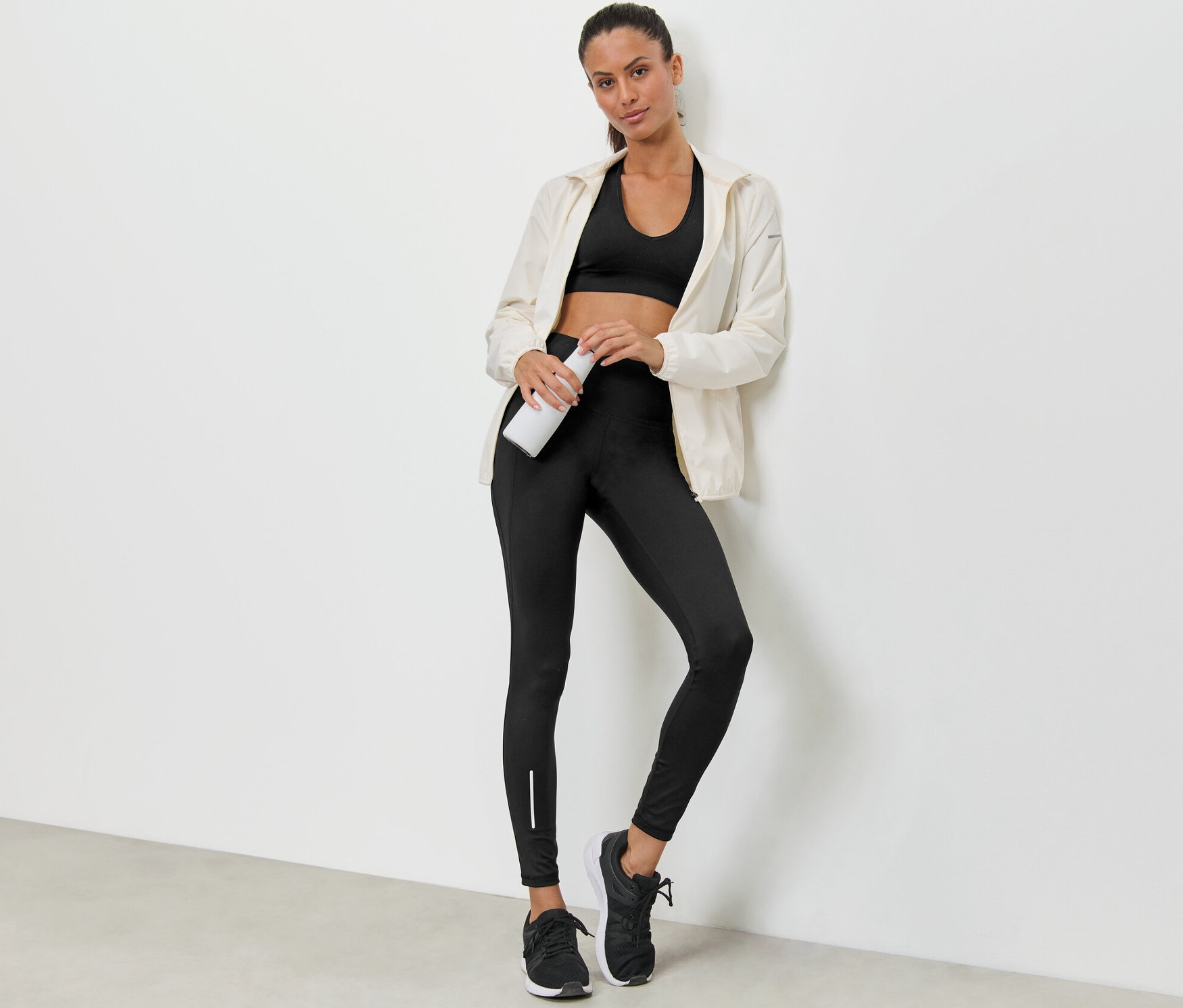 Eine Frau lehnt an einer Wand, hält eine Wasserflasche und trägt einen Sport-BH, eine Jacke und eine wasserabweisende Sporttight.