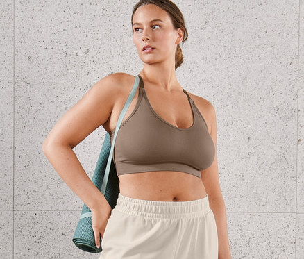 Frau trägt ein braunes Seamless-Sportbustier und eine extraweiche Sweathose. Eine Yogamatte ist über ihre Schulter gehängt.