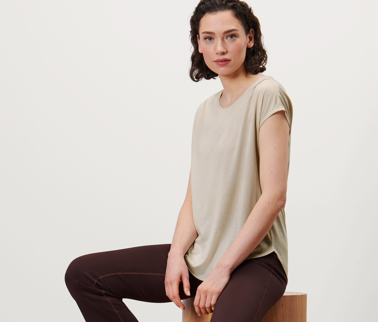 Frau sitzt auf Holzzylindern und trägt ein beiges Sportshirt und braune Hosen.