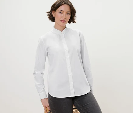 Eine Frau posiert auf einem Stuhl und trägt eine weiße Bluse mit Rüschendetail und graue Jeans.