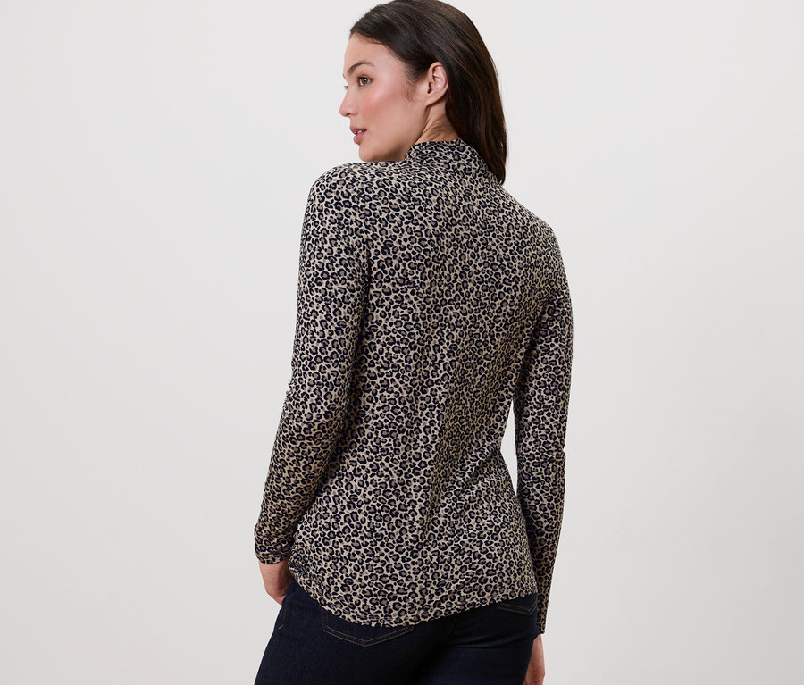 Eine Frau steht mit dem Rücken zur Kamera und trägt ein bedrucktes Blusenshirt mit Stehkragen und dunkle Jeans.