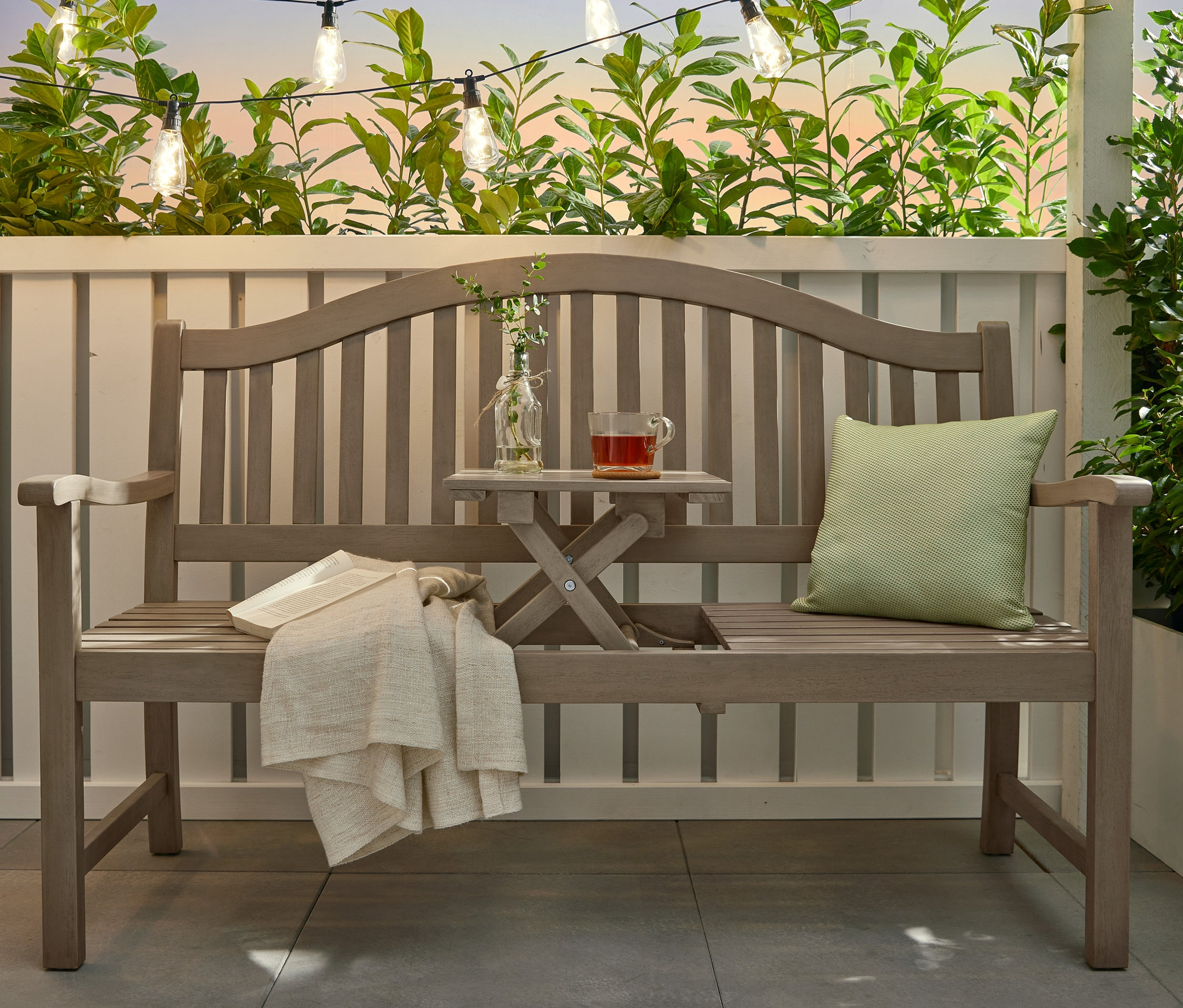 Graue Bank »Leira« mit hochklappbarem Tabletttisch mit Buch und Decke darauf. Über der Bank hängt eine LED-Filament-Lichterkette.