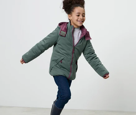 Lächelndes Mädchen in 2-in-1-Kinder-Softshell-und-Steppjacke mit Kapuze, Jeans und Gummistiefeln.