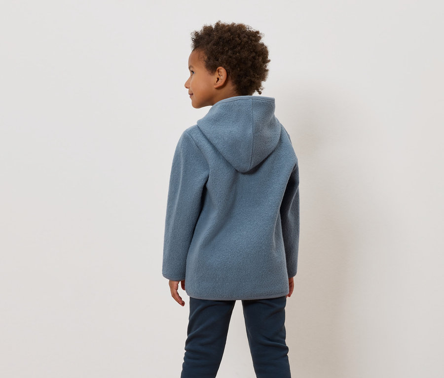 Ein Junge mit lockigem Haar trägt eine blaue Kapuzenjacke und dunkelblaue Hosen.